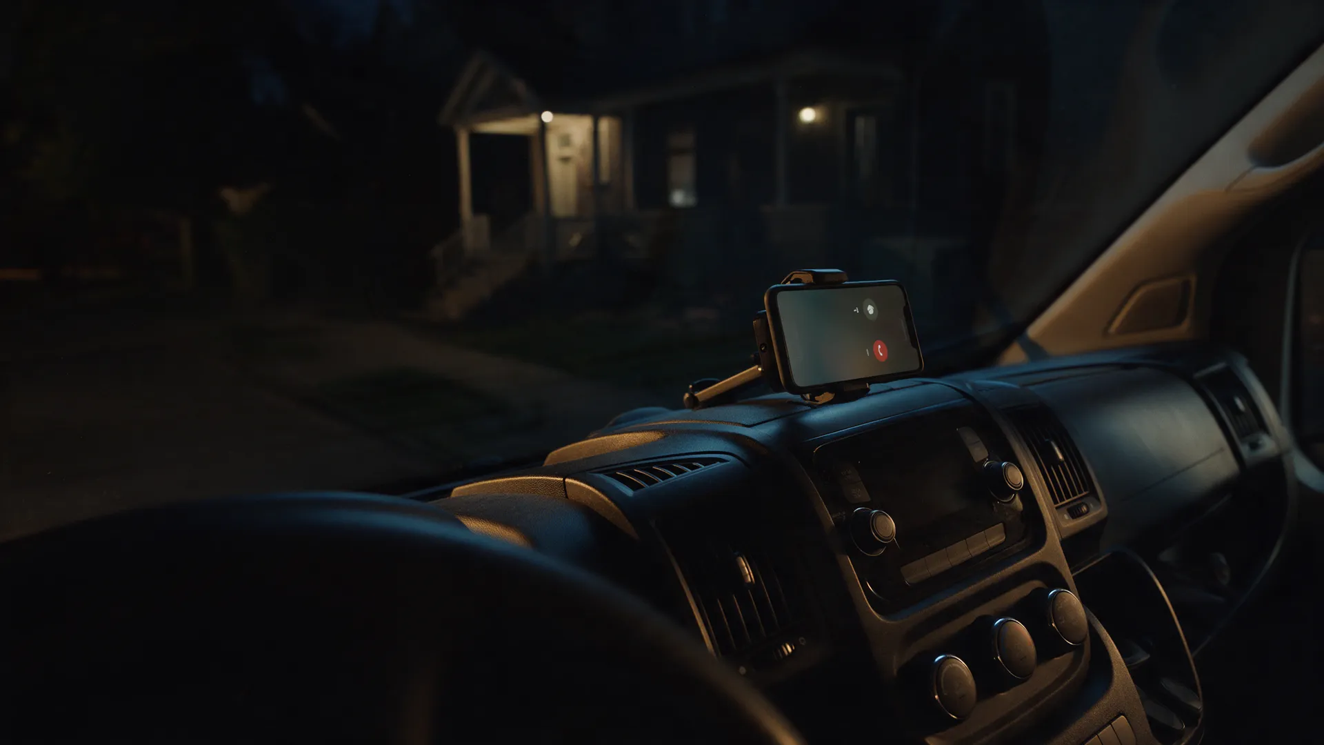 A phone in a dashboard mount lighting up with a call at night, a lit porch beyond the windscreen.